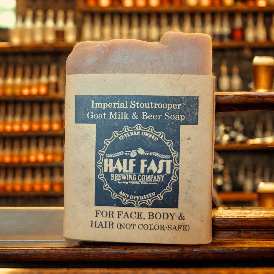 A bar of 'Imperial Stoutrooper' goat milk and beer soap resting on a wooden surface with shelves of various items in the background. The soap bar is packaged in a brown paper wrapper with branding details.