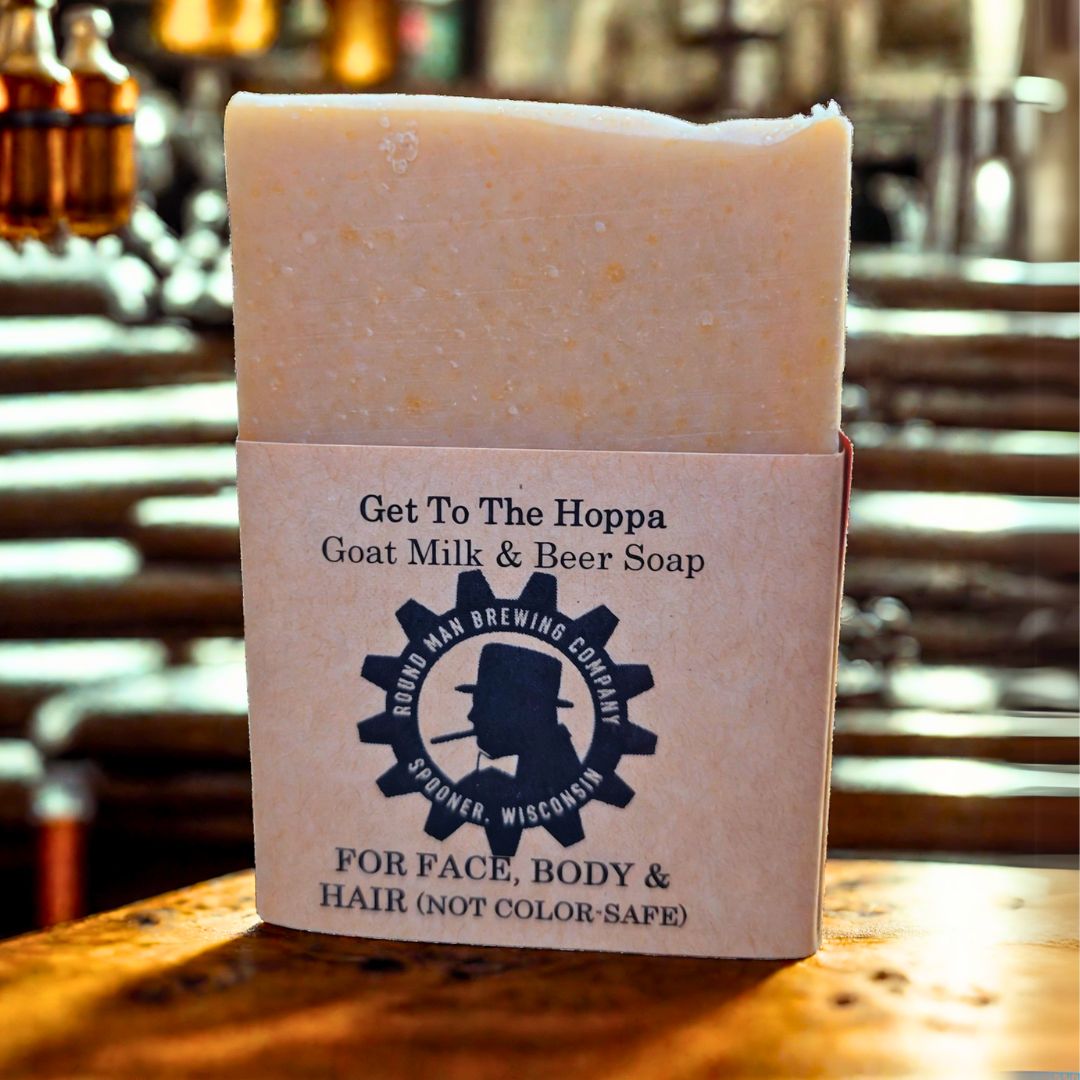 A bar of 'Get to the Hoppa' Goat Milk & Beer Soap with packaging that includes the product name and a note saying 'FOR FACE, BODY & HAIR (NOT COLOR-SAFE)'.
