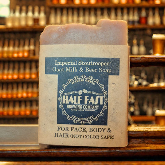 A bar of 'Imperial Stoutrooper' goat milk and beer soap resting on a wooden surface with shelves of various items in the background. The soap bar is packaged in a brown paper wrapper with branding details.