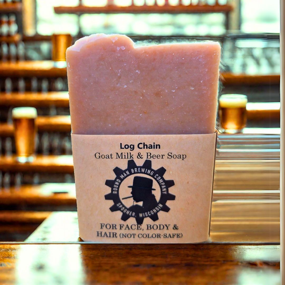 A bar of 'Log Chain' Goat Milk & Beer Soap with a beige color, resting on a wooden surface, with a label that includes the product name and purposes for face, body, and hair.